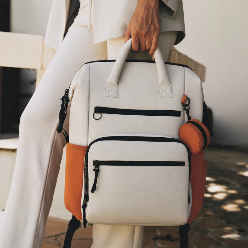 Frau in heller Kleidung trägt den Wickelrucksack in Creme-Beige und Apricot am oberen Tragegriff